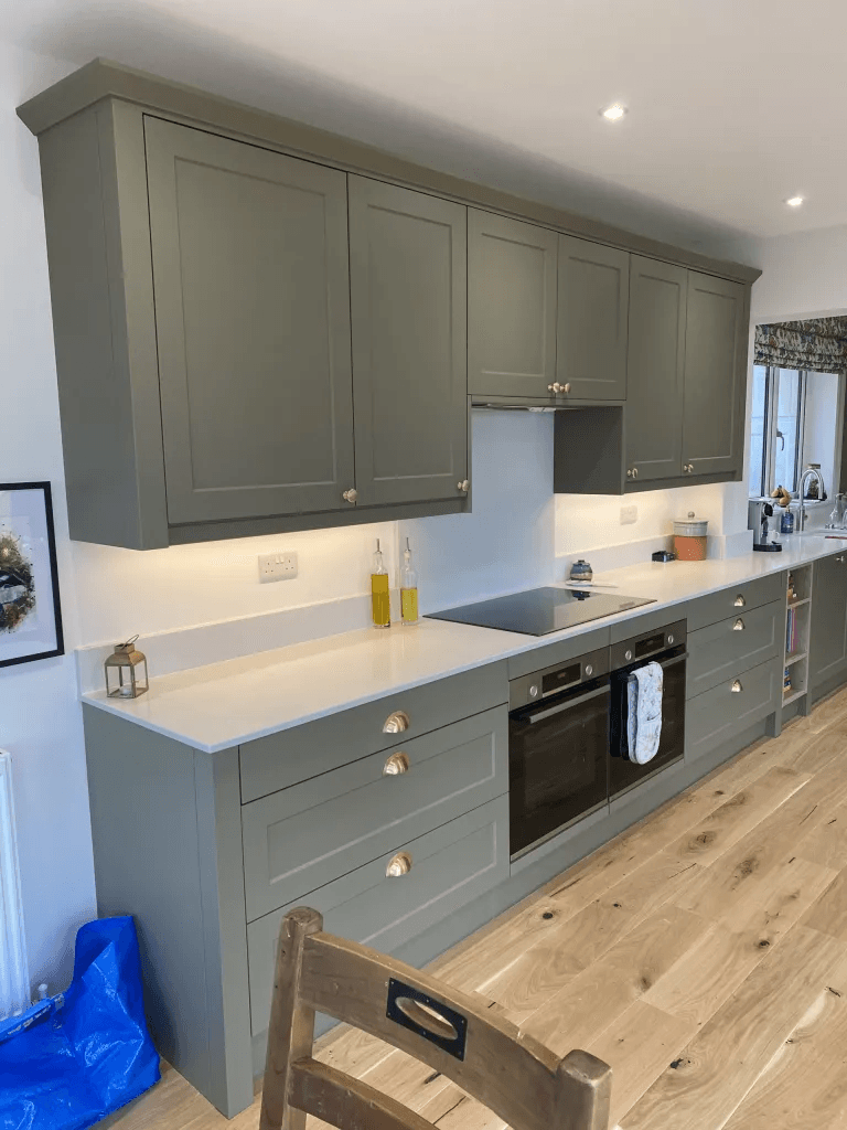 A kitchen with green shaker cabinets, brass handles, white worktops and integrated ovens
